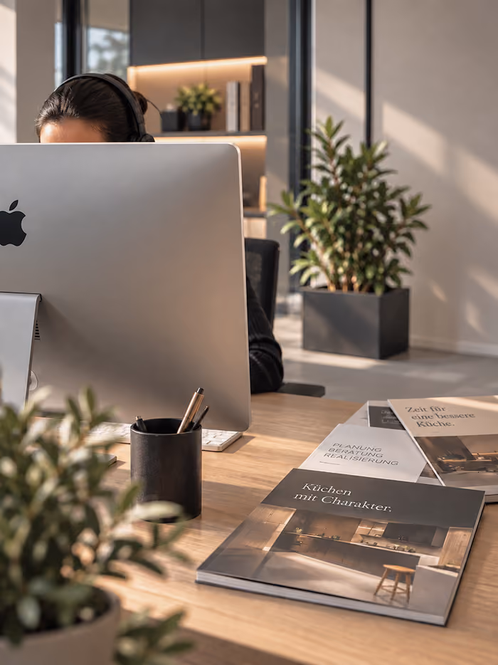 Frau mit Headset sitzt hinter einem Apple-Computer an einem hellen Holztisch mit Katalogen über Küchen auf der rechten Seite, im Hintergrund Pflanzen und Bücherregal.