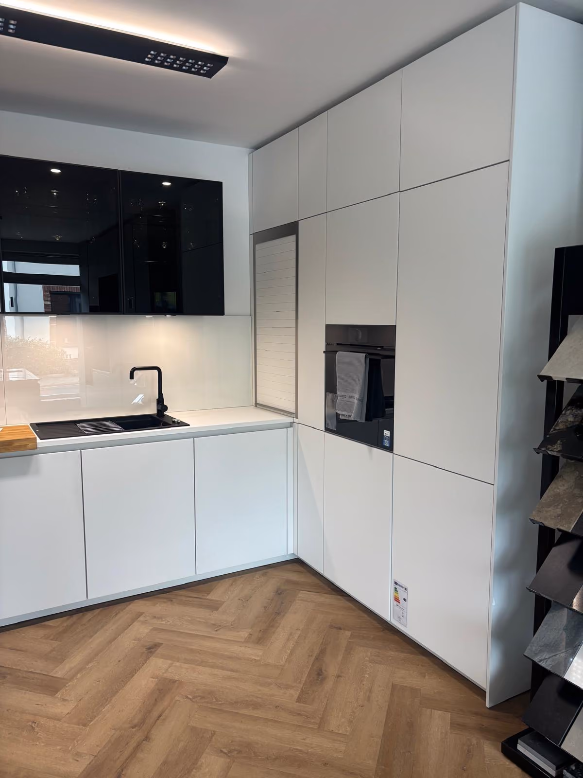 Moderne Küche mit weißer und schwarzer Einbauküche, integriertem Backofen, schwarzer Spüle und Wasserhahn sowie einem Holzfußboden im Fischgrätmuster.