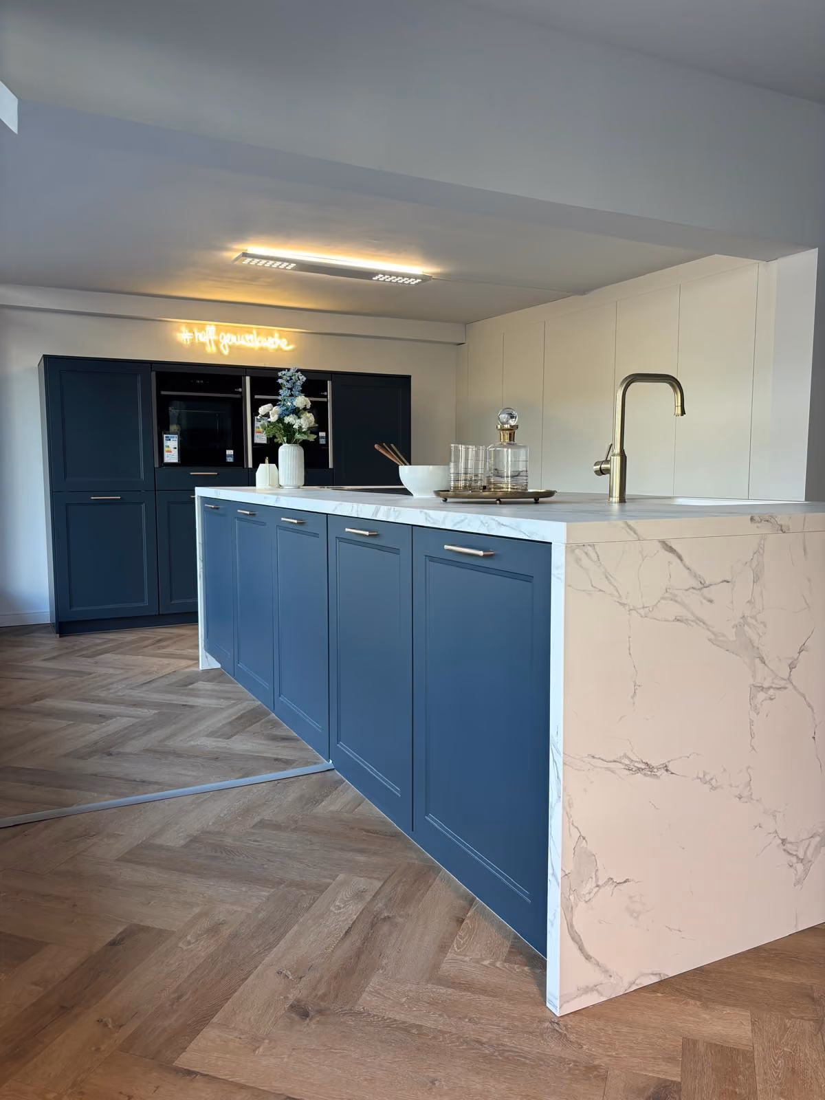 Moderne Küche mit blau lackierten Unterschränken und einer weißen Marmor-Arbeitsplatte mit grauen Adern, einer goldfarbenen Wasserarmatur und einem großen Blumenarrangement im Hintergrund neben eingebauten schwarzen Küchengeräten unter einer warmen Deckenbeleuchtung.