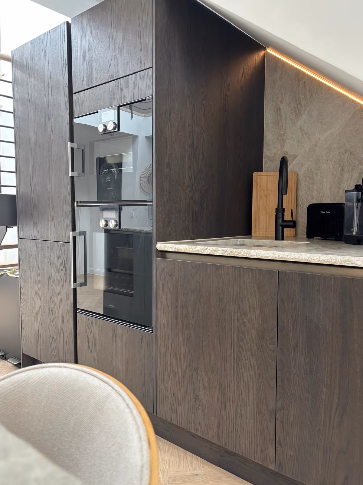 Moderne Küchenschränke aus dunklem Holz mit eingebautem Doppelbackofen, hellem Stein-Arbeitsplattenbereich, schwarzem Wasserhahn und einem Schneidebrett an der Wand unter einer geneigten Decke mit indirekter Beleuchtung.