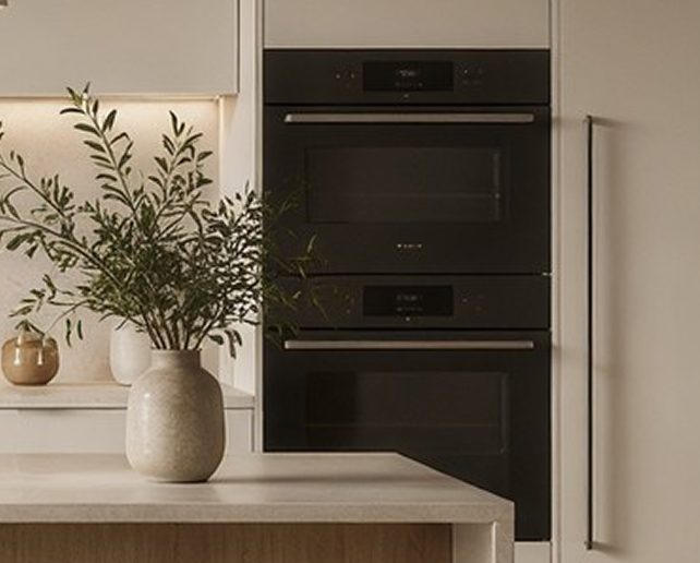 Moderne Küche mit eingebauten schwarzen Backöfen und einer hellen Arbeitsplatte, auf der eine Vase mit grünen Zweigen steht.