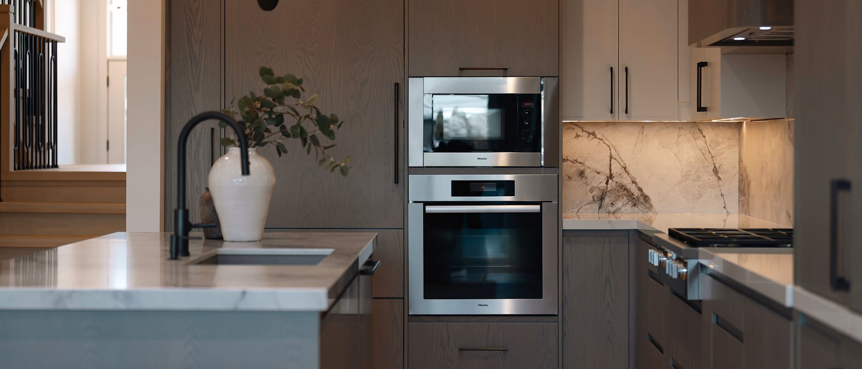 Alt-Text: „Moderne Küche mit dunklen Holzfronten, Kücheninsel, Backofen und beleuchteter Naturstein-Rückwand.