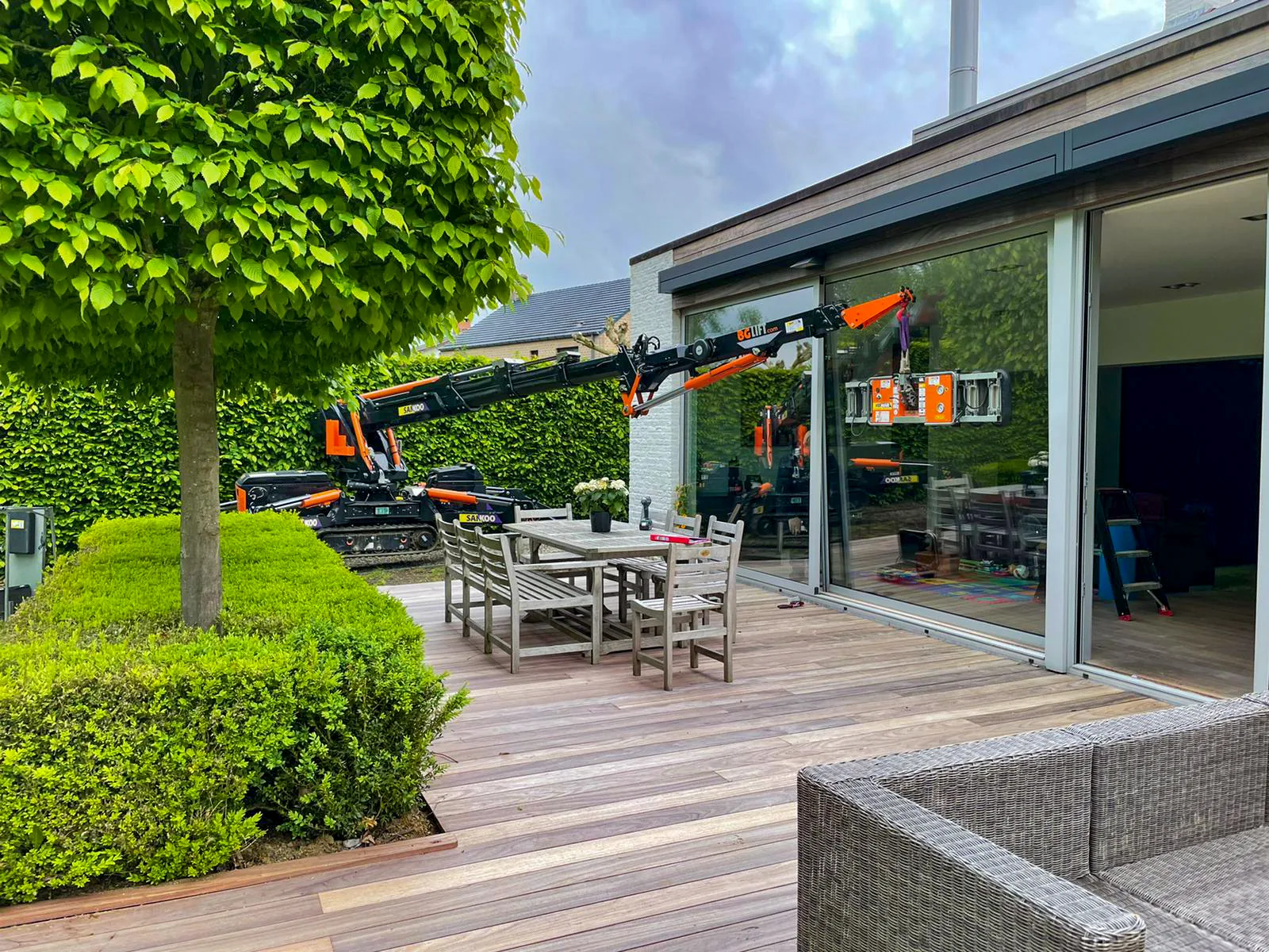 Orange tracked spider crane lifting equipment beside a house with glass doors and deck.