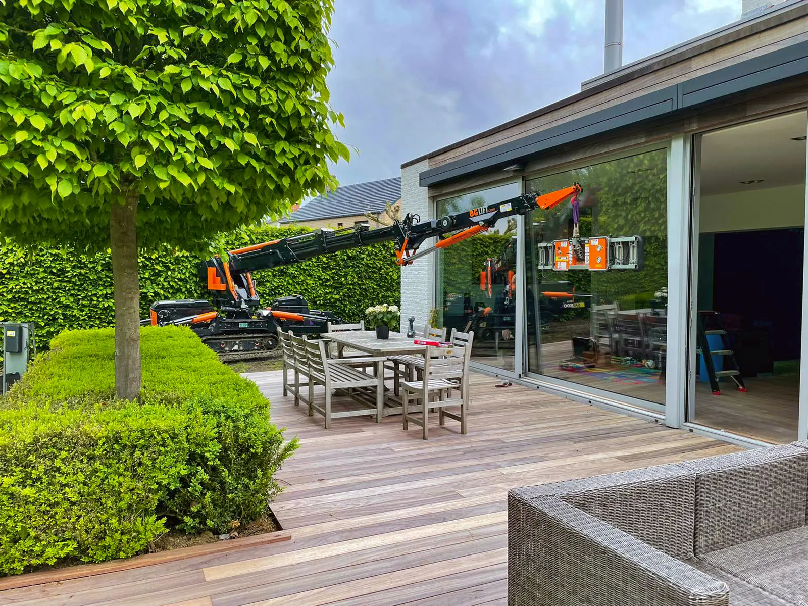 Outdoor wooden deck with a dining table and chairs near a modern house with large sliding glass doors, where a compact crane is lifting a glass panel for installation.