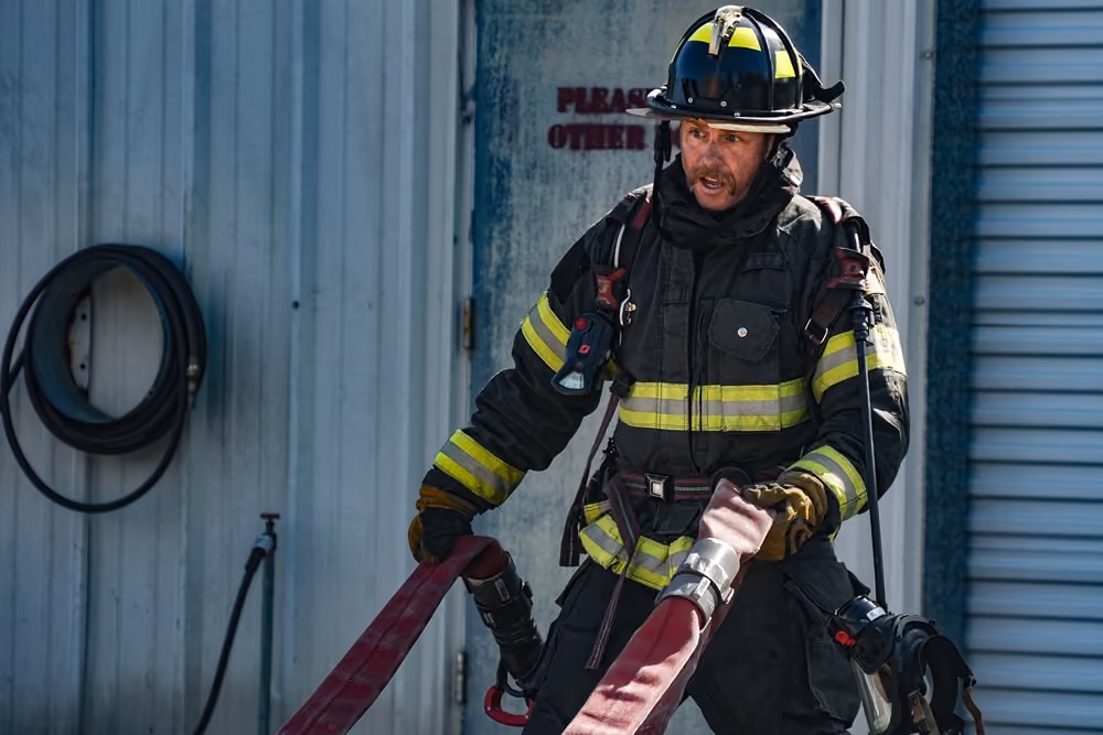Firefighter in helmet and turnout gear carrying a hose in front of a metal roll-up door