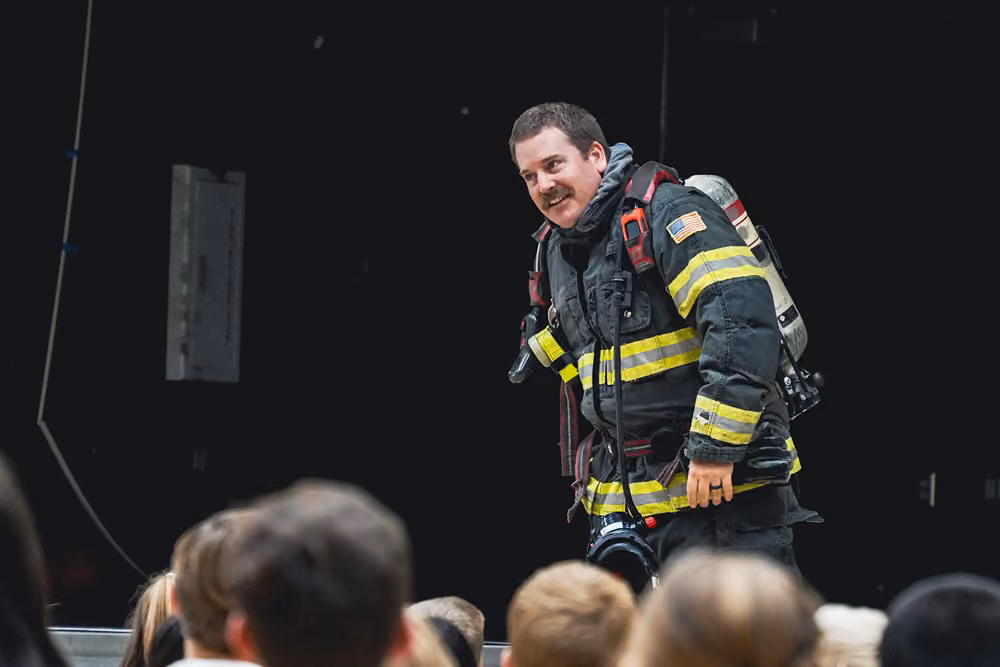 Firefighter in turnout gear and SCBA walking on stage in front of a seated audience