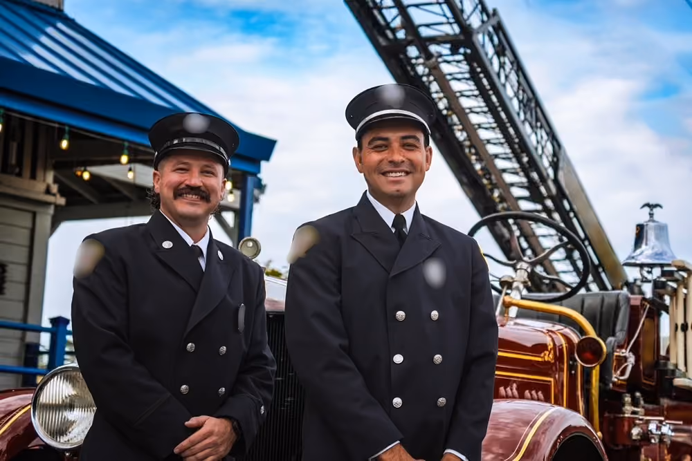 Two firefighters in dress uniform standing in front of a vintage fire truck
