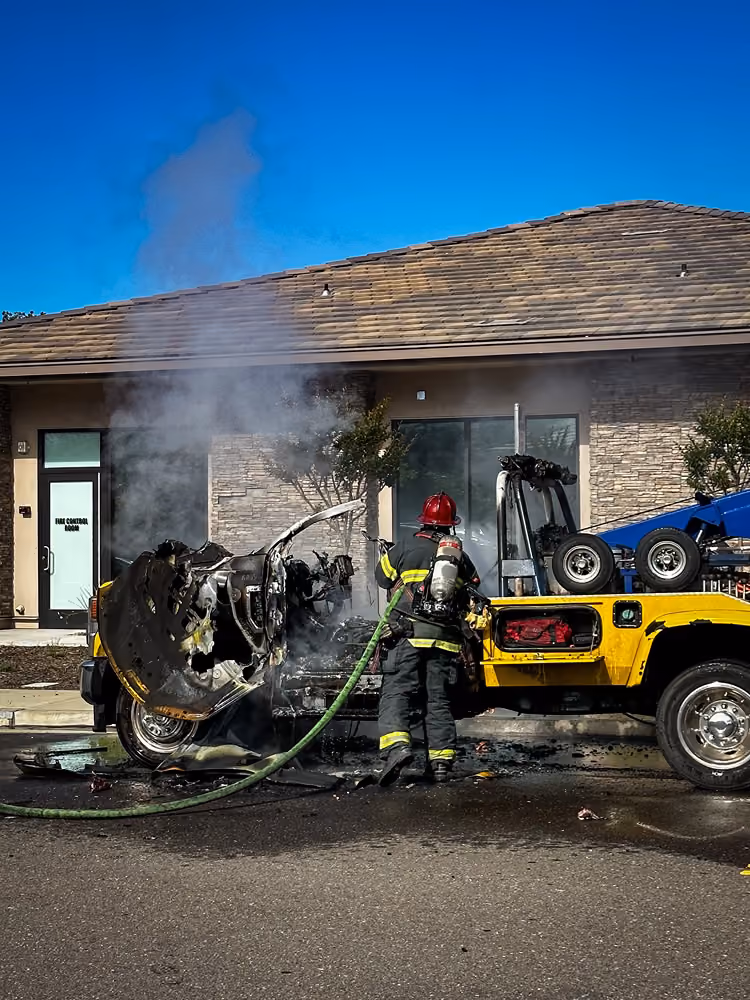 Firefighter in turnout gear extinguishing a burned vehicle fire in a parking lot, with a tow truck nearby