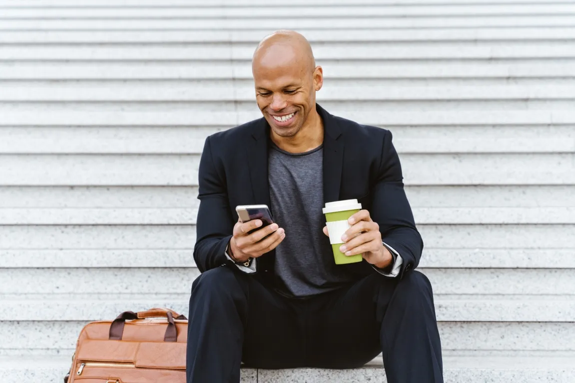 A professionally dressed man sitting on building steps, engaged in a video call on his phone while holding a cup of coffee.
