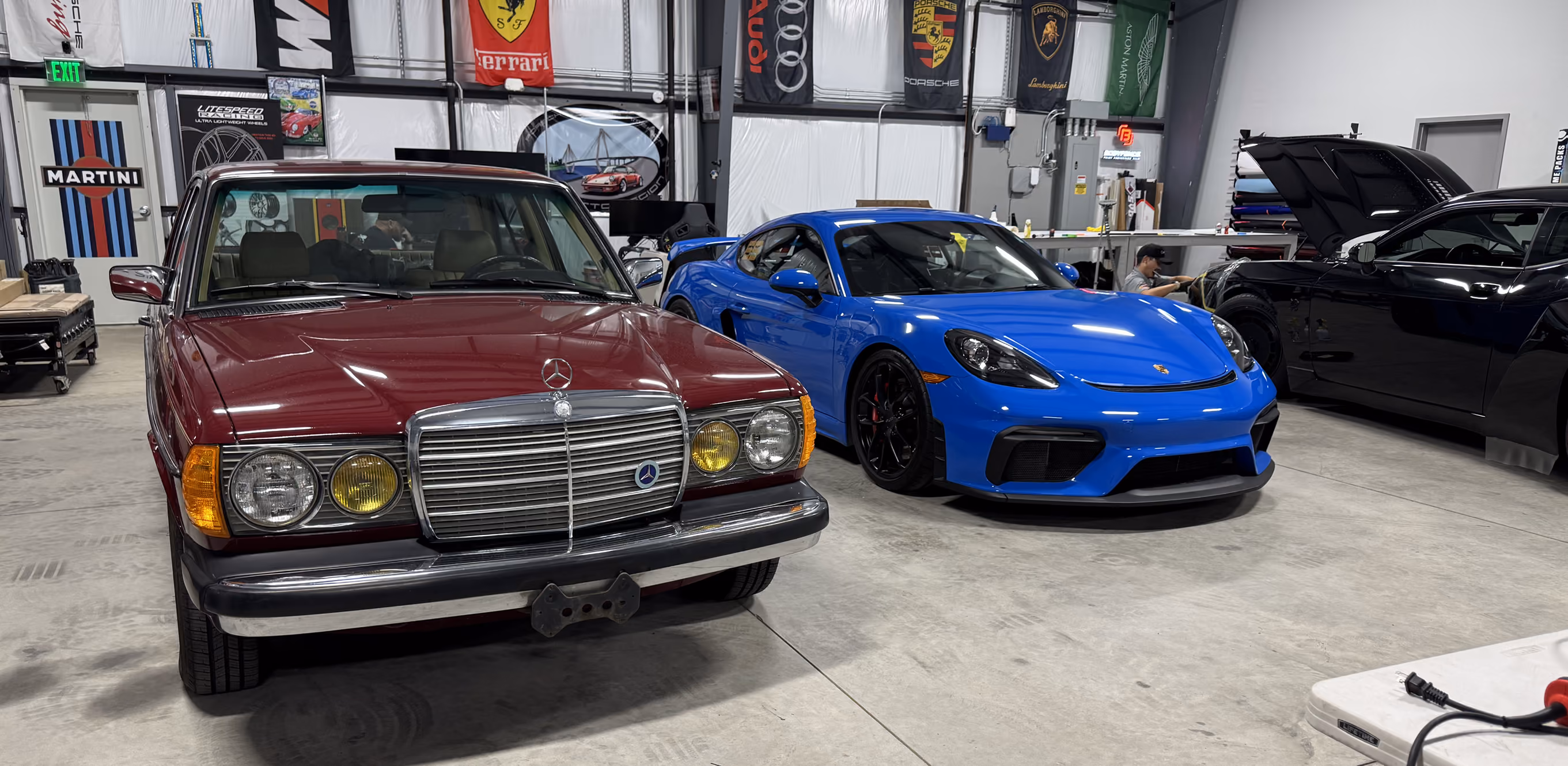 Vintage maroon Mercedes and modern blue Porsche sports cars parked inside a garage with car brand flags.