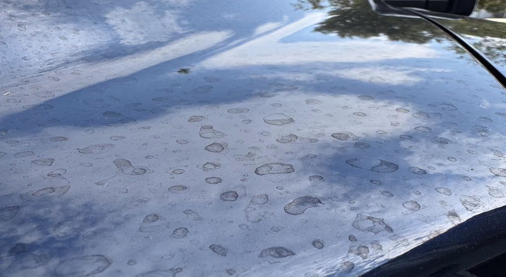 Car hood covered with water spots and light reflections of sky and clouds.