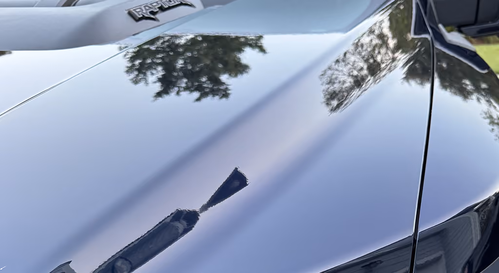 Glossy black car hood with a Raptor badge and reflections of trees and a lamp post.