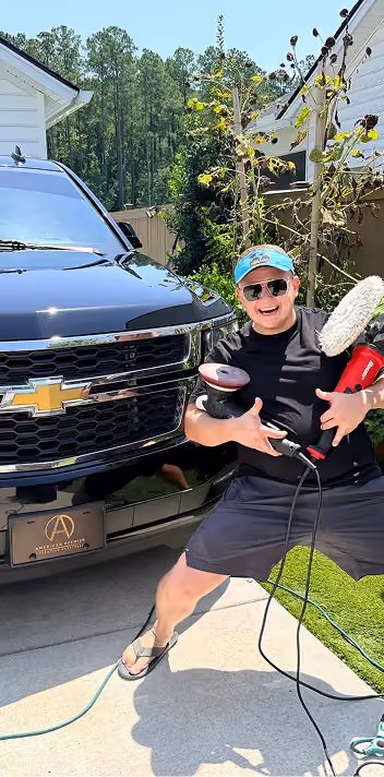 Man wearing sunglasses, black T-shirt, shorts, and a blue cap poses energetically in front of a black Chevrolet truck, holding car polishing equipment on a driveway with greenery and houses in the background.