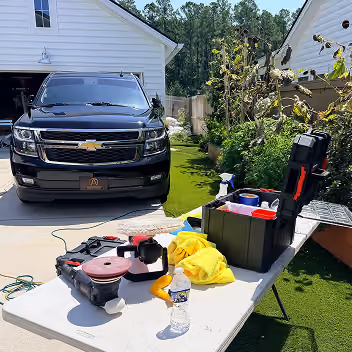 Black Chevrolet SUV parked in a driveway next to a white house, with a folding table in front holding various tools, a toolbox, yellow gloves, and a water bottle, with green grass and trees in the background.