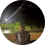 Person wearing a black leather jacket sitting outdoors at night facing a misty, empty basketball court with bright floodlights and trees in the background.