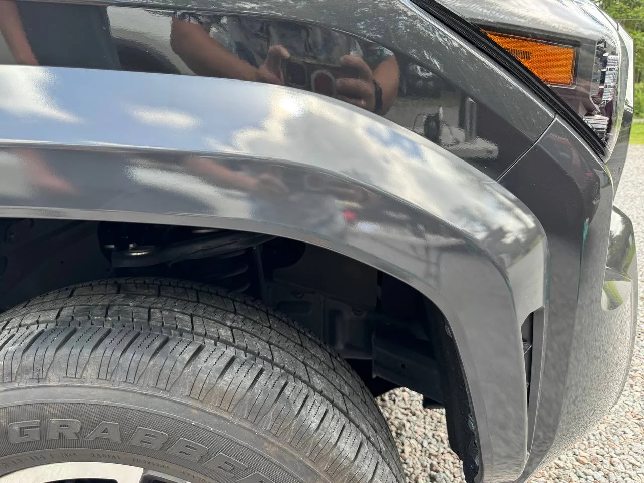 Close-up of the front left wheel and fender of a black vehicle on a gravel surface, showing tire tread and part of the headlight and indicator light.