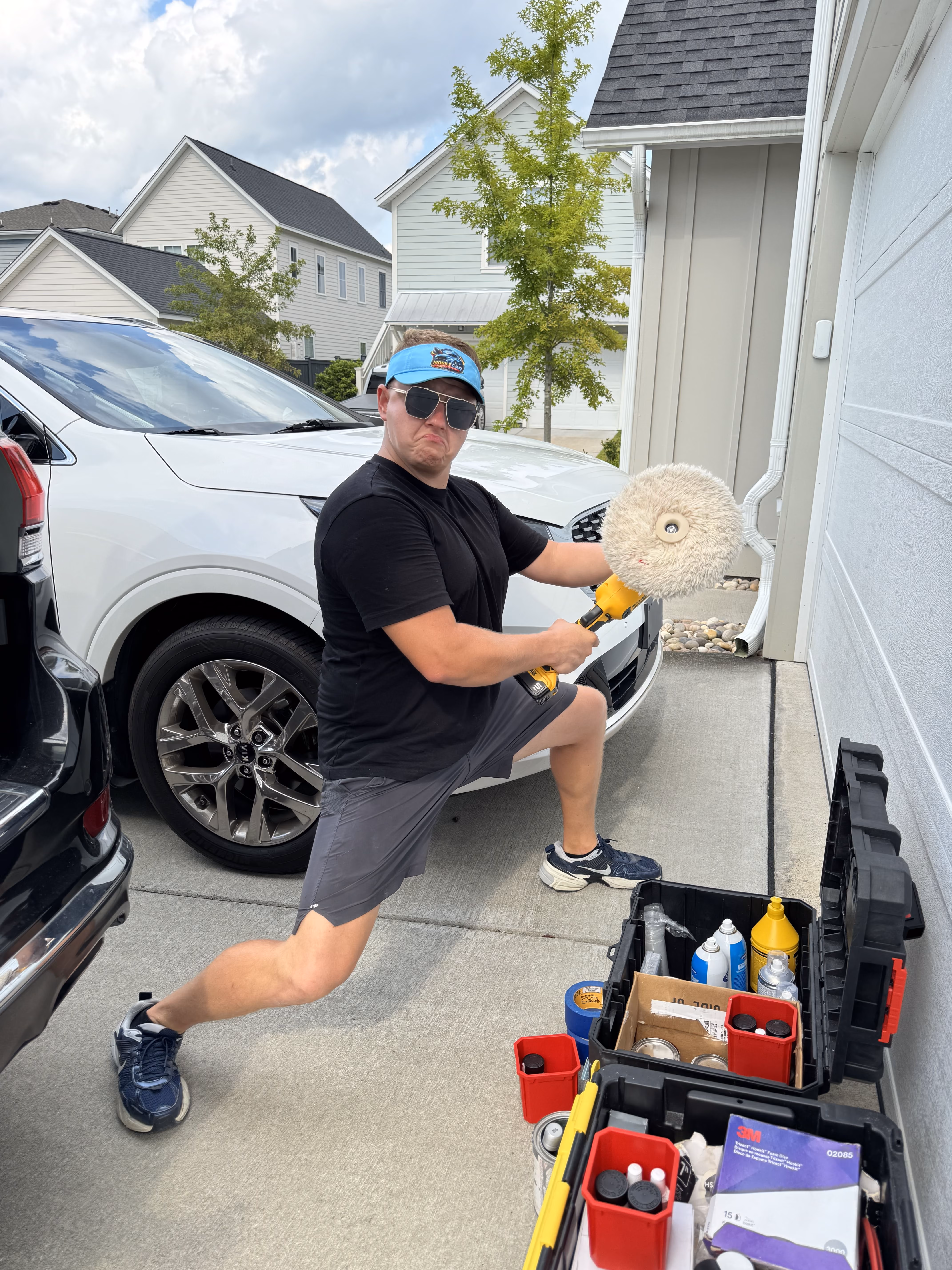 A man wearing a black t-shirt, gray shorts, blue shoes, sunglasses, and a blue baseball cap poses in a wide stance on a driveway holding a large buffing tool, with open toolboxes and car care products nearby and white cars parked in the background.