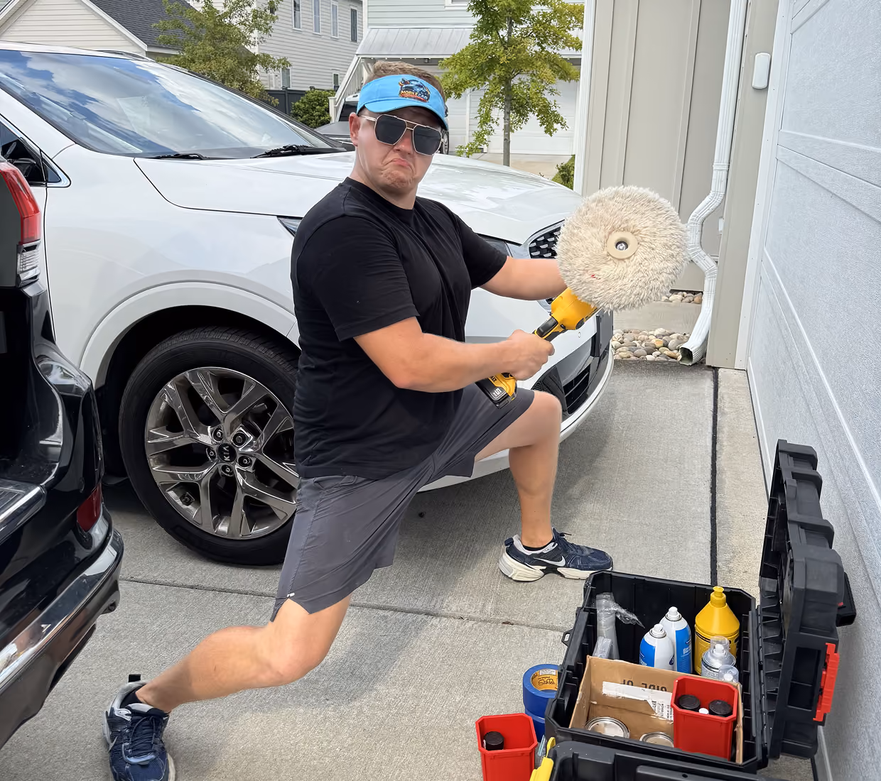 Man in black shirt, gray shorts, blue visor, and sunglasses playfully posing with a car buffer in a driveway between a black and a white car next to an open black toolbox filled with cleaning supplies.