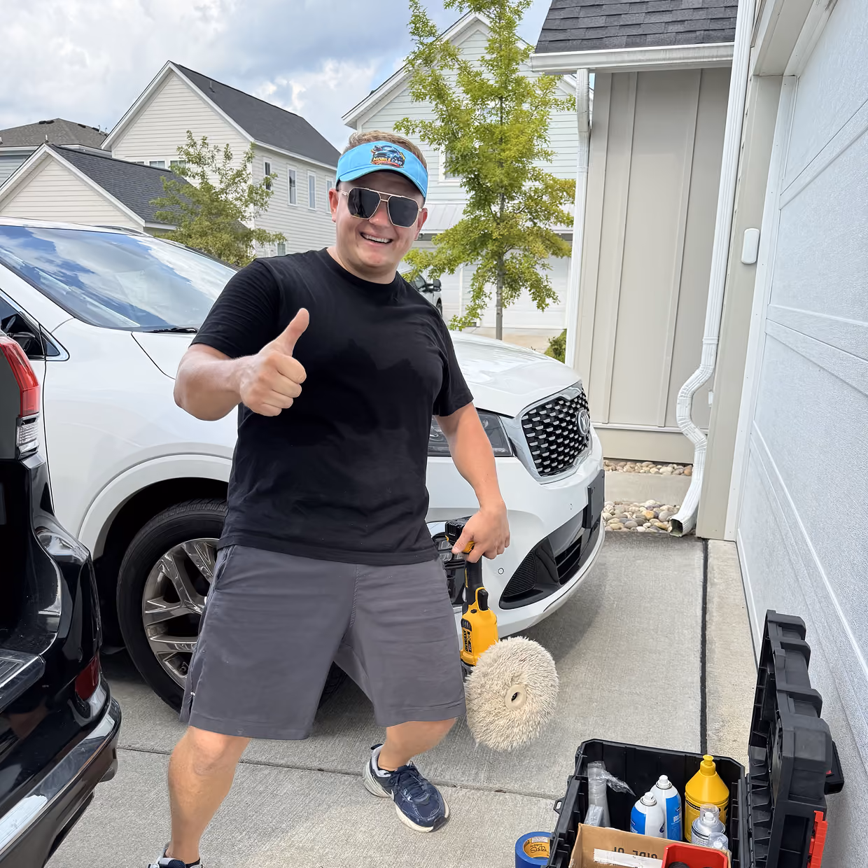 Smiling man wearing sunglasses, a blue visor, black t-shirt, and gray shorts stands on a driveway between black and white cars, holding a car detailing buffing tool and giving a thumbs-up, next to an open toolbox with car cleaning supplies.