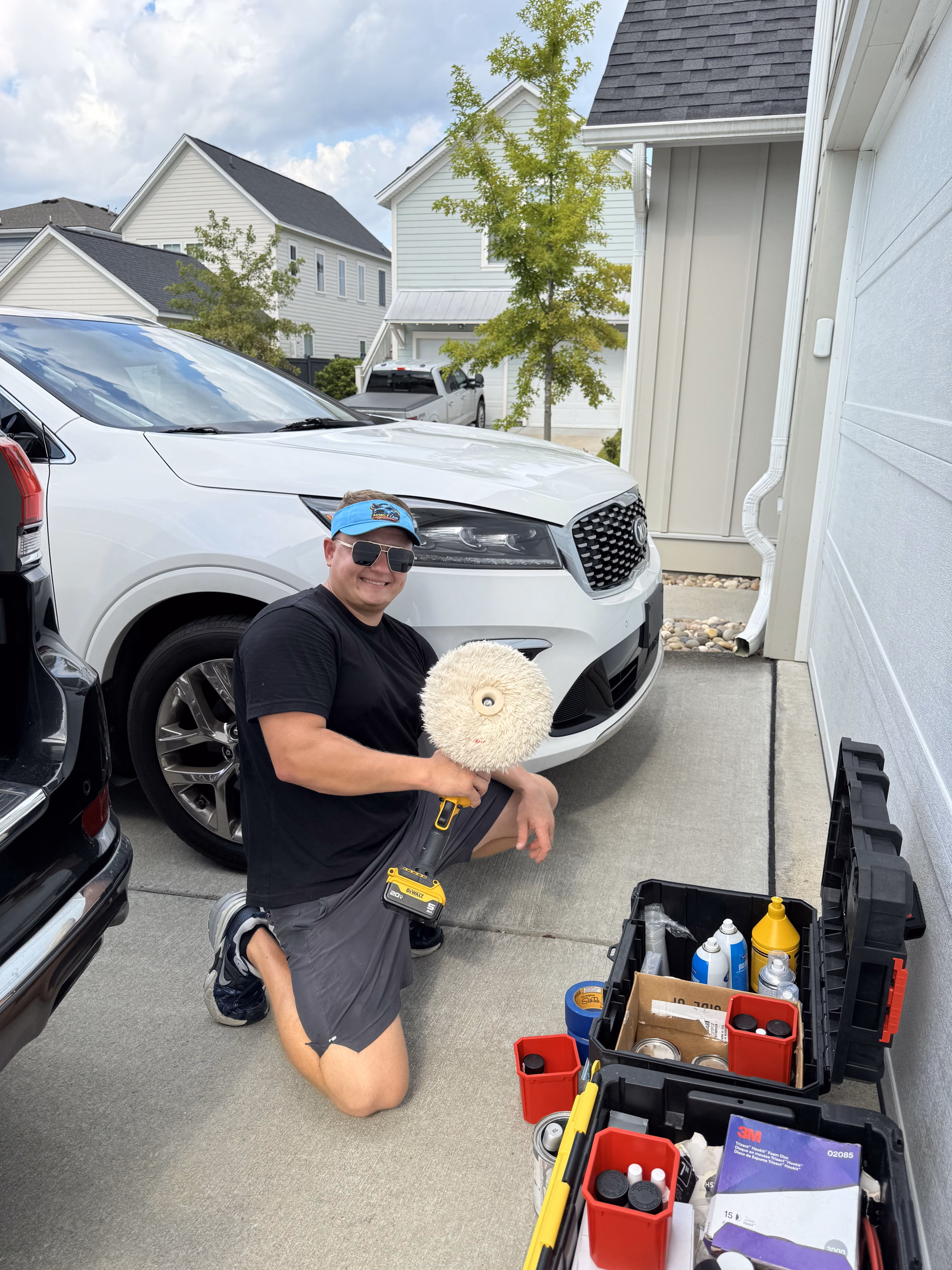 A man wearing sunglasses, a blue visor, black t-shirt, and gray shorts kneels on a driveway holding a vehicle buffer polisher while smiling at the camera, with a white SUV and toolboxes containing car care products nearby.