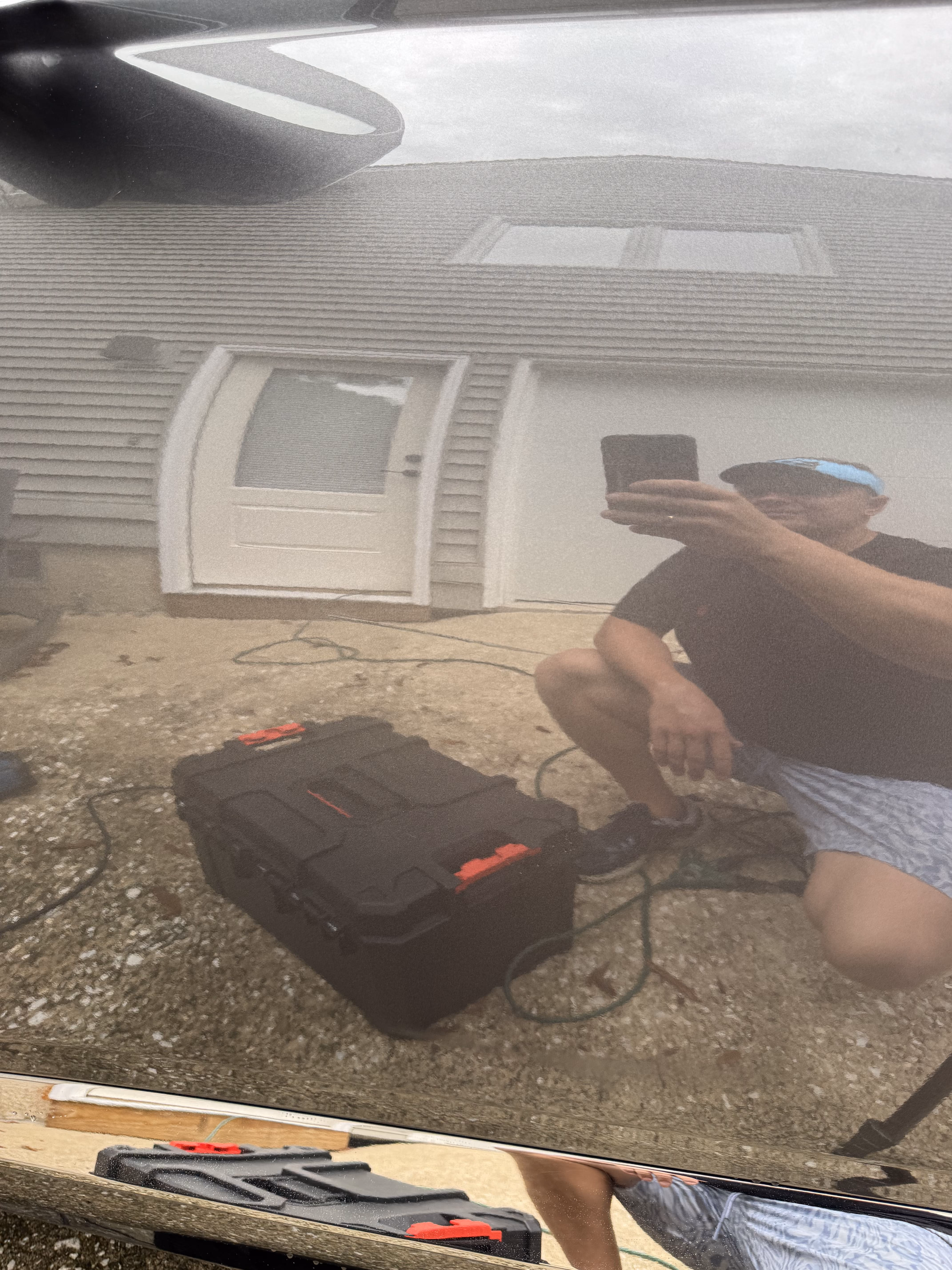 Reflection of a man taking a photo with a smartphone, kneeling next to a black toolbox with red latches on a gravel surface in front of a white house with a door and window.