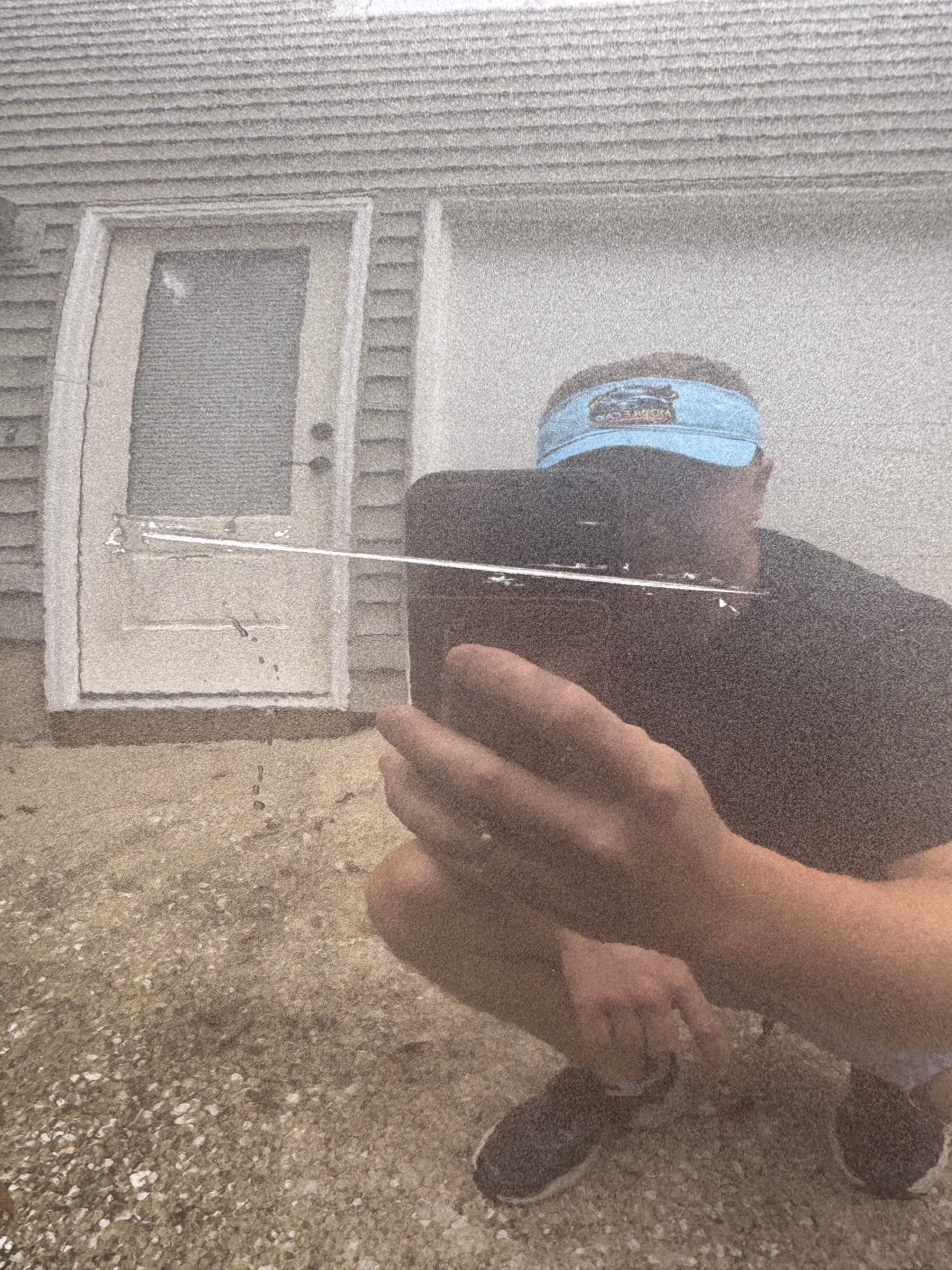 Reflection of a person wearing a blue visor and black shirt, crouching and holding a phone, seen on a glossy surface with a long scratch across it, with a beige house door and siding in the background.