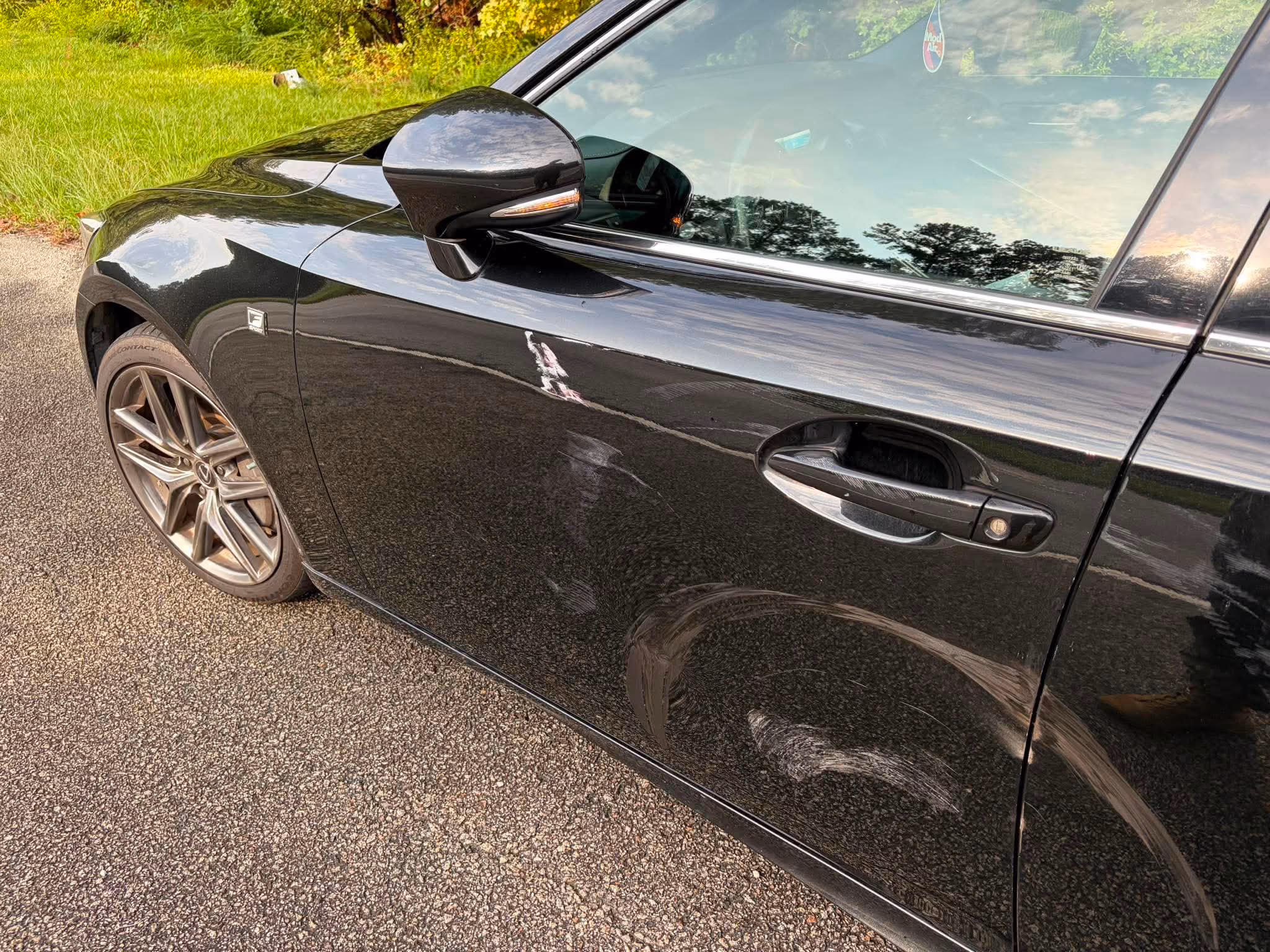 Close-up of the driver's side of a black car with visible scratches and scuff marks on the door near the handle and lower panel, parked on a gravel surface with grass and trees in the background.