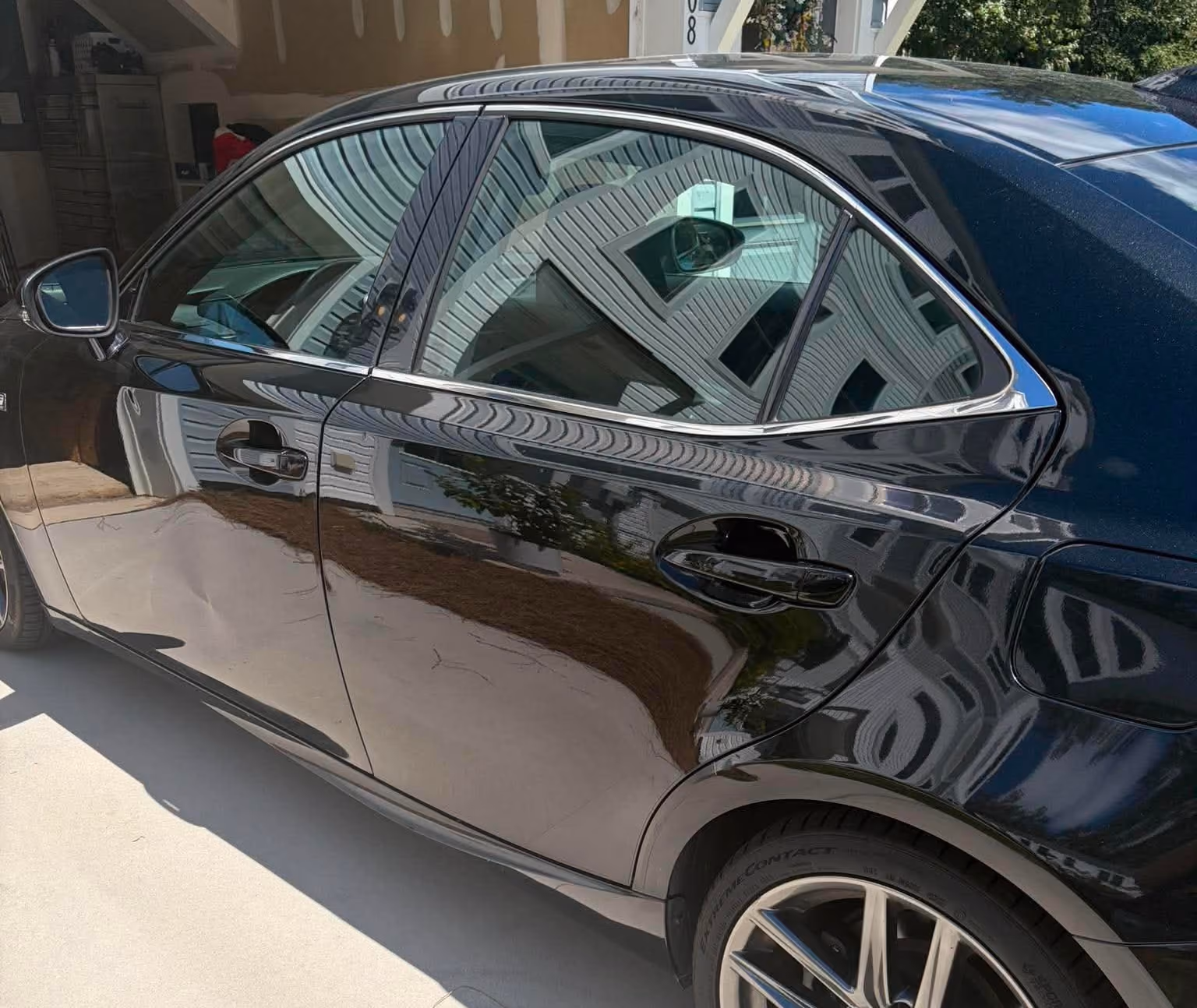 Close-up of the side of a black car with a large dent on the rear door panel, parked on a driveway in front of a garage, with reflections of nearby house siding on the car surface.