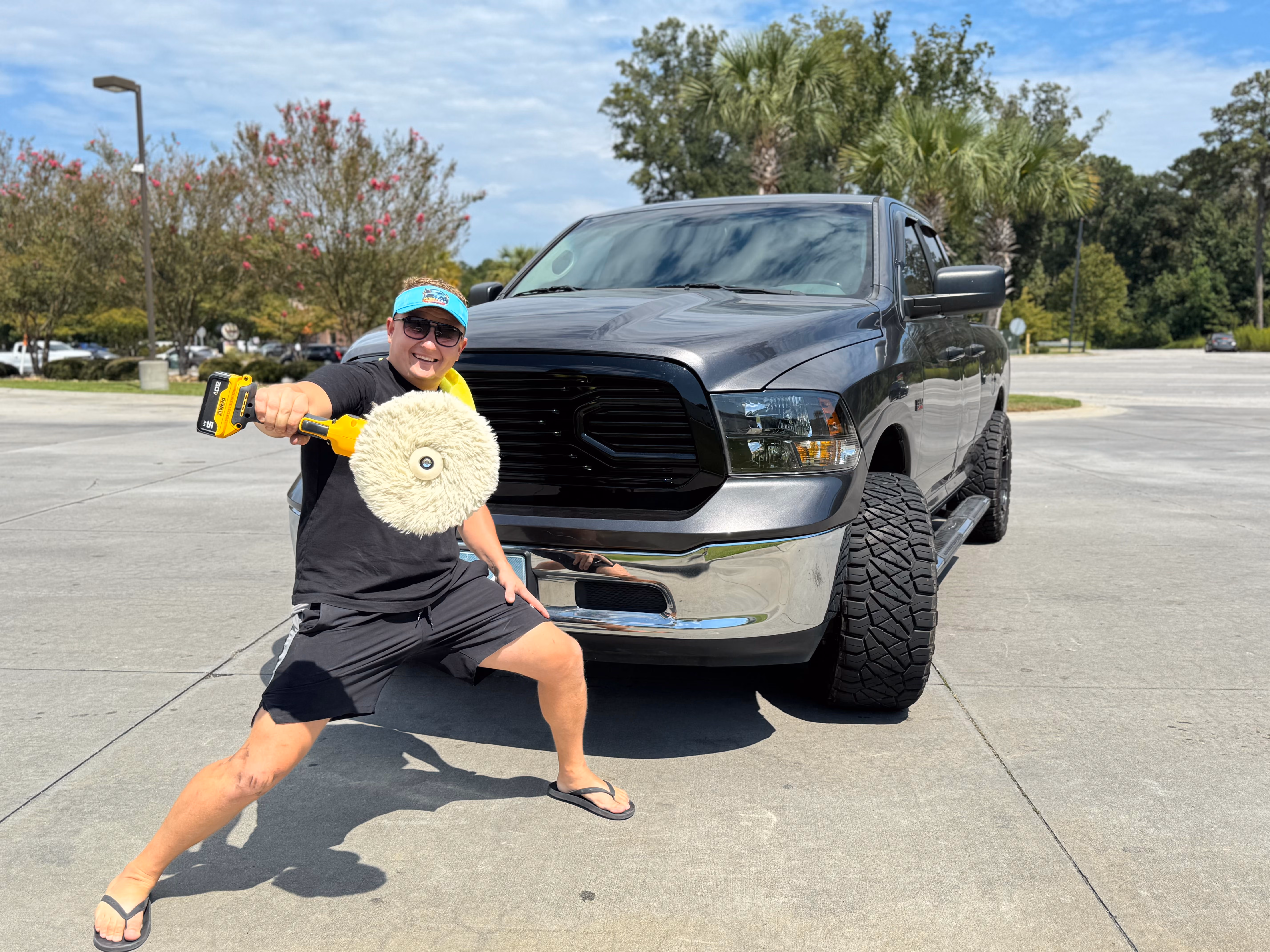 A man wearing sunglasses, a black shirt, shorts, flip-flops, and a teal visor holds a polishing machine with a buffing pad towards the camera while standing in front of a large gray pickup truck in a parking lot on a sunny day.