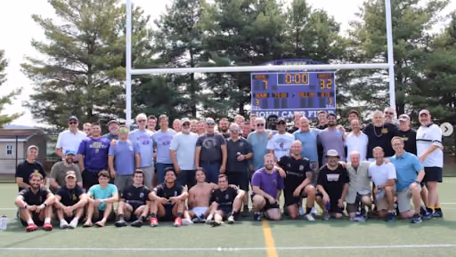 Large group of men posing together on a sports field in front of a scoreboard that shows the score 7 to 32 with time 0:00, with trees in the background.