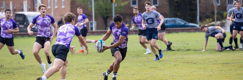 A rugby player in a purple and black uniform running with the ball while two opposing players in gray uniforms approach on a grassy field during a match.