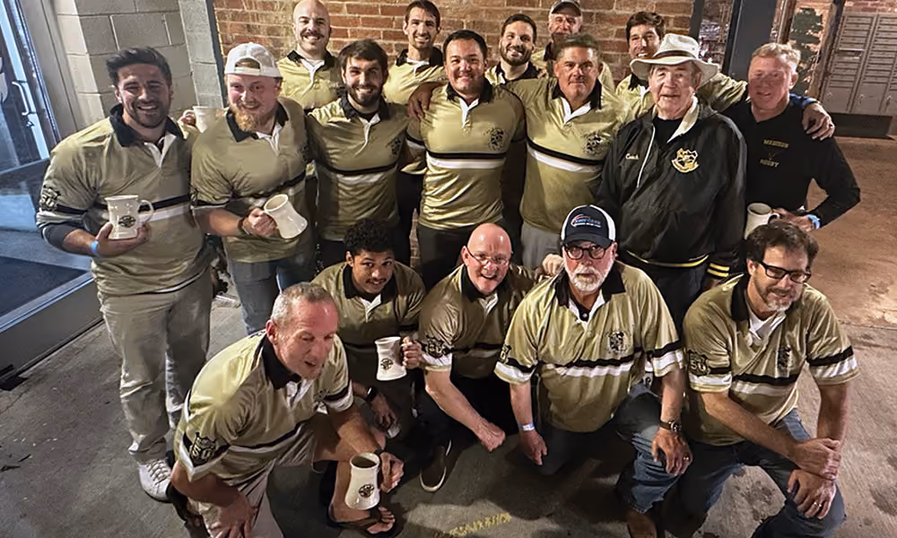 Group of fifteen men wearing matching beige and black rugby shirts posing indoors with brick wall background, some holding white mugs, smiling and crouching or standing together.