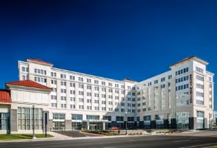 Large, multi-story white building with many windows under a clear deep blue sky, situated along a street with manicured grass and small bushes.