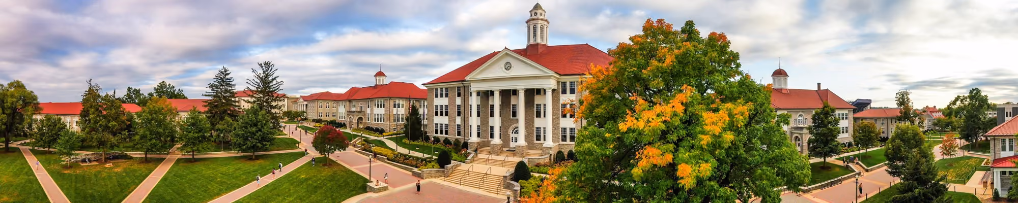Panoramic view of a university campus with a prominent building featuring tall white columns and a red roof, surrounded by walkways, green lawns, trees, and additional buildings under a partly cloudy sky.