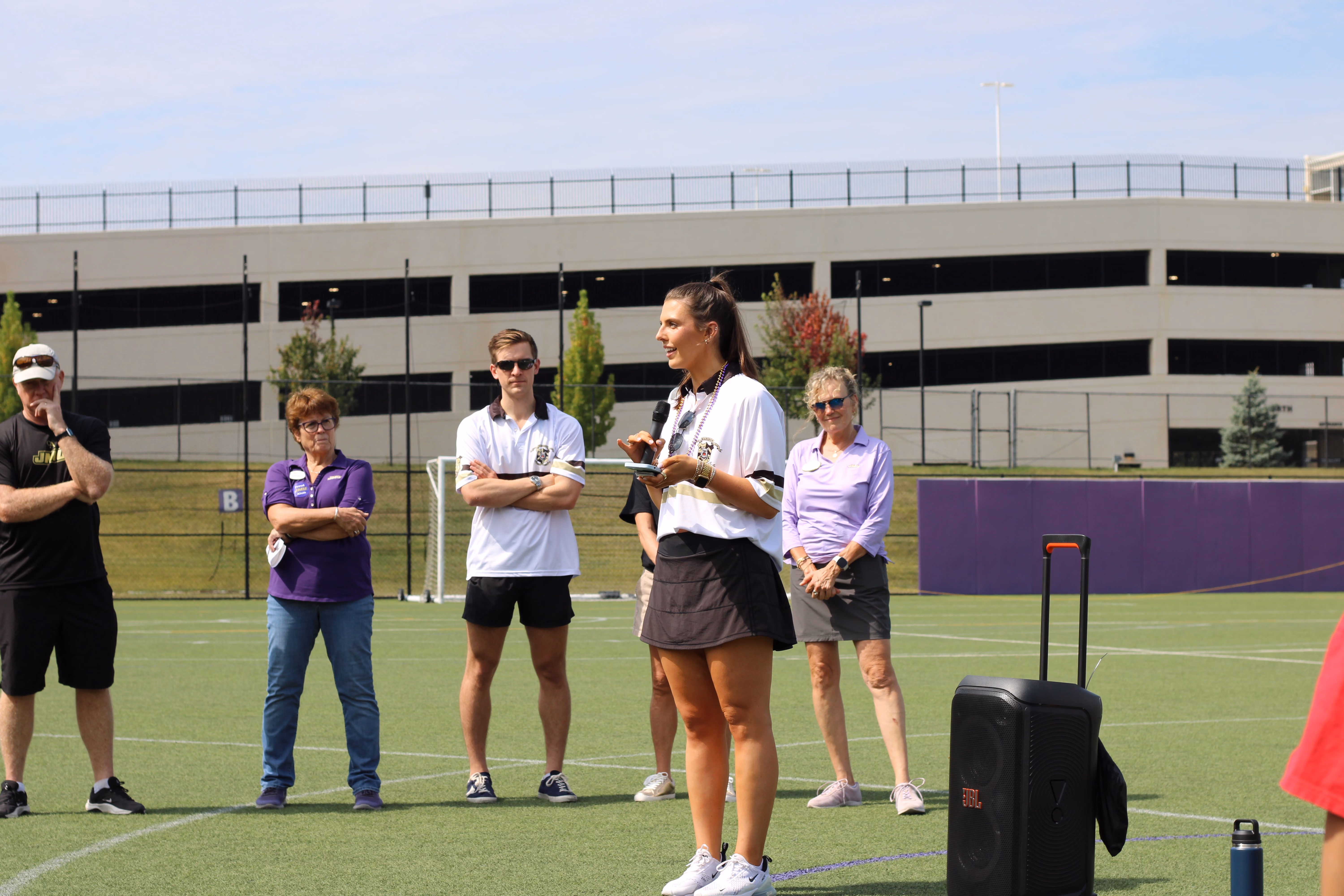 Woman in sports attire holding a microphone and speaking on a synthetic turf field, with four individuals standing and listening in the background near a parking garage.