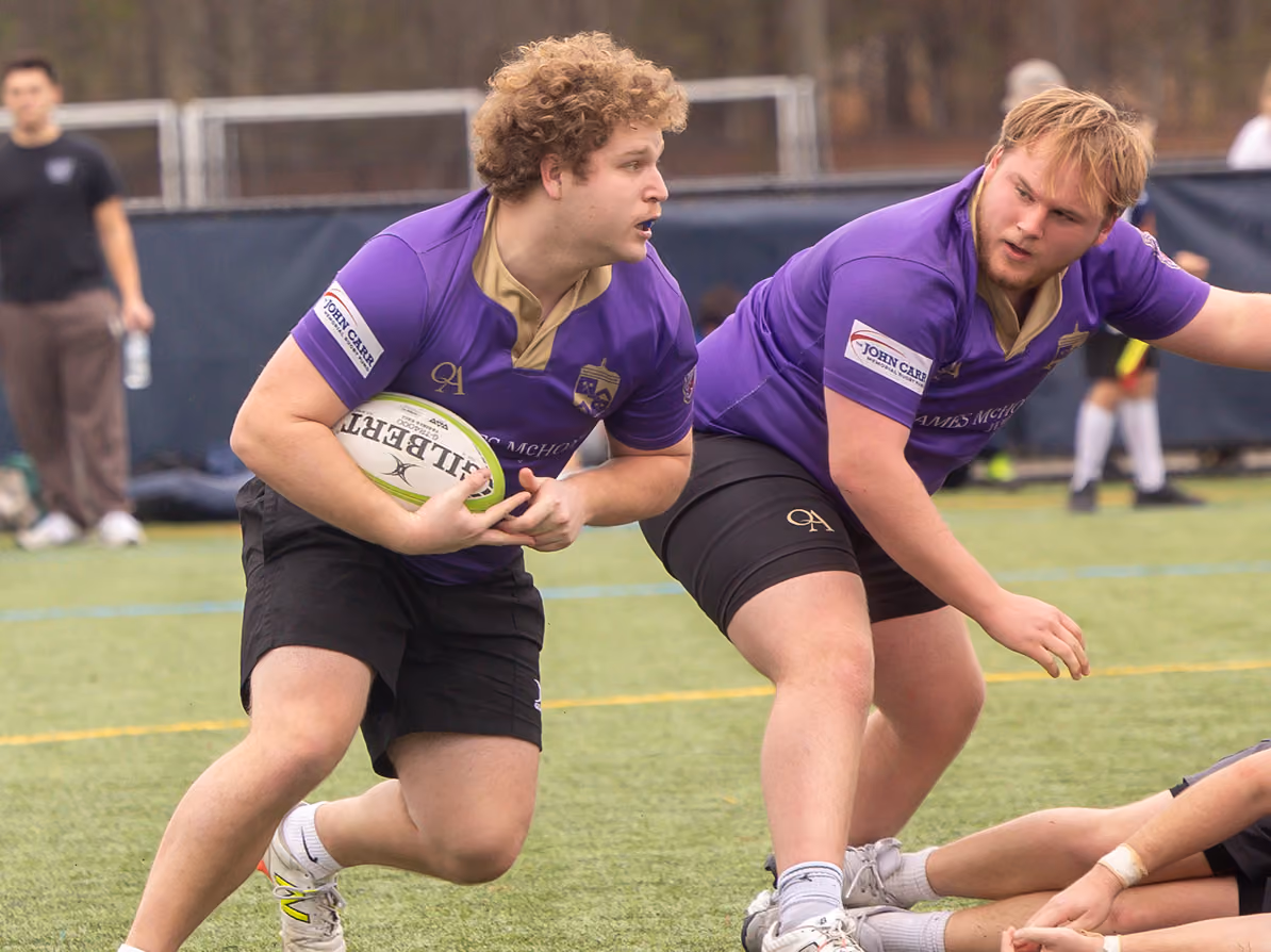Two young male rugby players in purple jerseys and black shorts during a game on a grassy field, one holding a rugby ball and preparing to pass it while the other braces nearby.