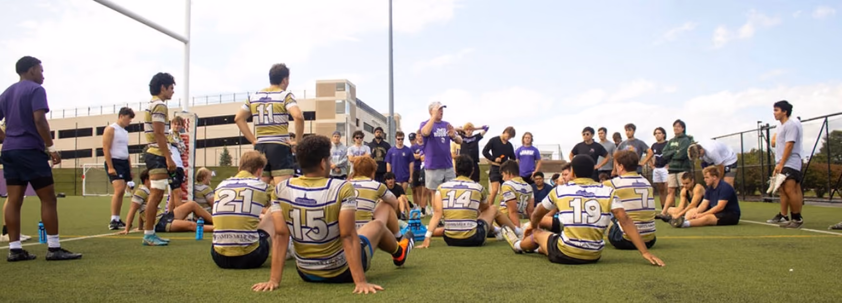 A rugby team in striped yellow and purple jerseys sitting and standing on a grass field, listening to a coach in a purple shirt giving instructions, with other players and spectators in the background.