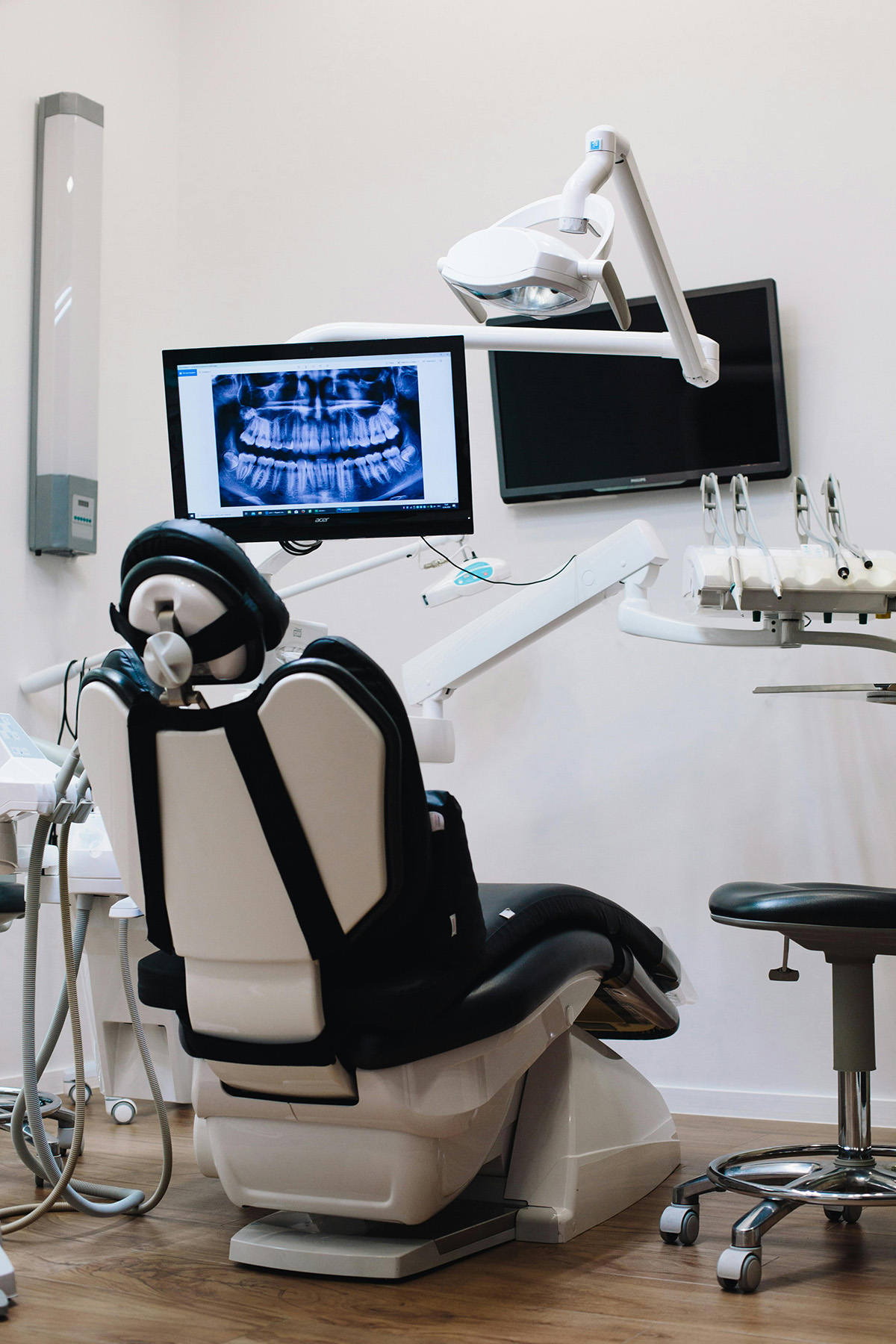 Empty dental treatment room with a chair, overhead light, and a monitor displaying a dental X-ray
