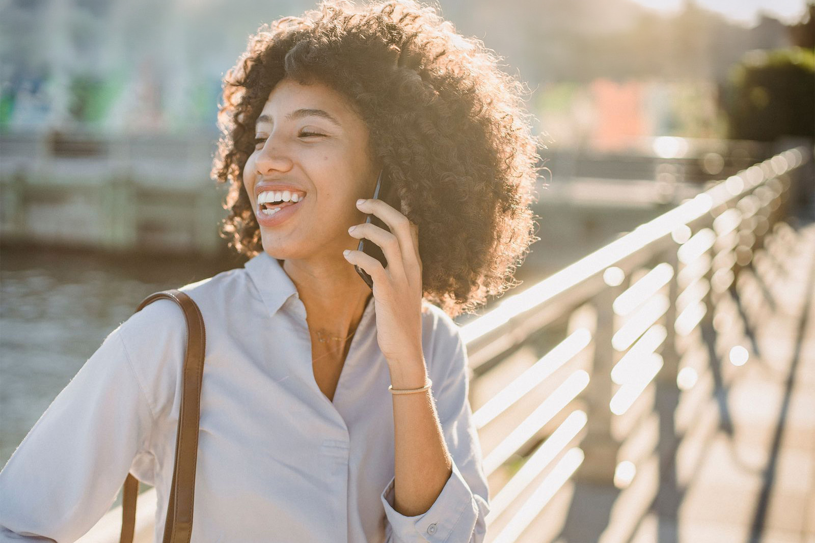 Smiling woman with curly hair laughing while talking on her phone outdoors near a waterfront
