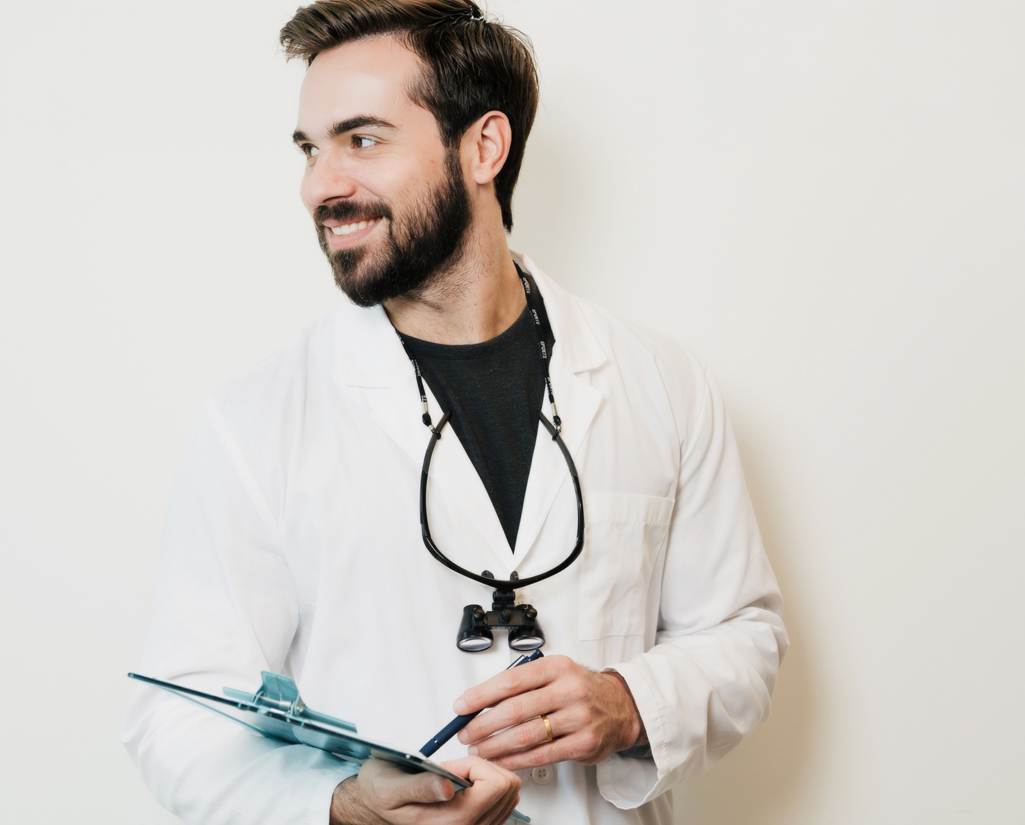 Smiling dentist in a white coat with dental loupes around his neck, holding a clipboard and pen