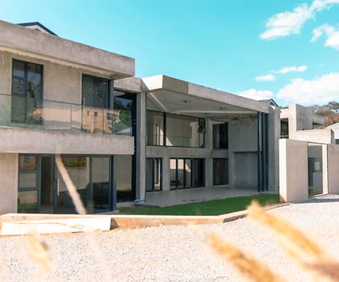 Modern two-story concrete building with large windows, glass railings, and a flat roof under a bright blue sky with scattered clouds.
