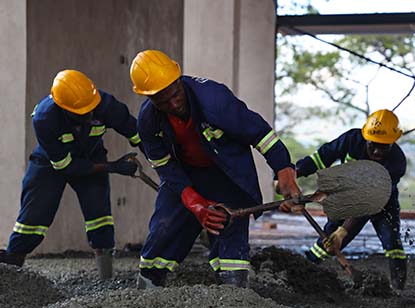Three construction workers wearing yellow helmets and blue work uniforms are spreading wet concrete indoors using tools.