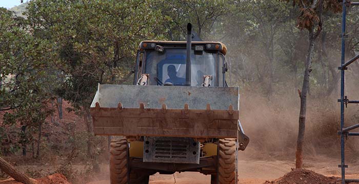 Front view of a bulldozer moving dirt on a forested construction site with dust rising around it and a driver visible inside the cabin.