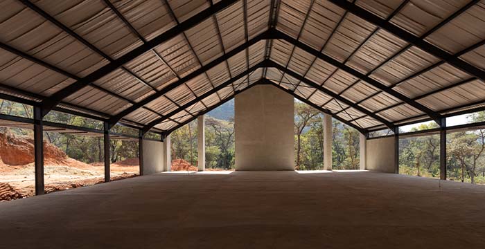 Interior view of a large, open construction building with a peaked metal roof and concrete floor, surrounded by natural trees and a dirt landscape visible through the open sides.