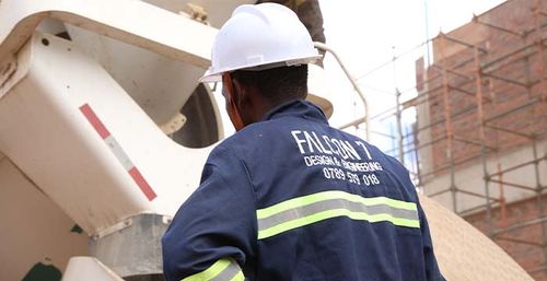 Construction worker wearing a white hard hat and navy blue jacket with reflective stripes operating machinery on a building site with scaffolding.