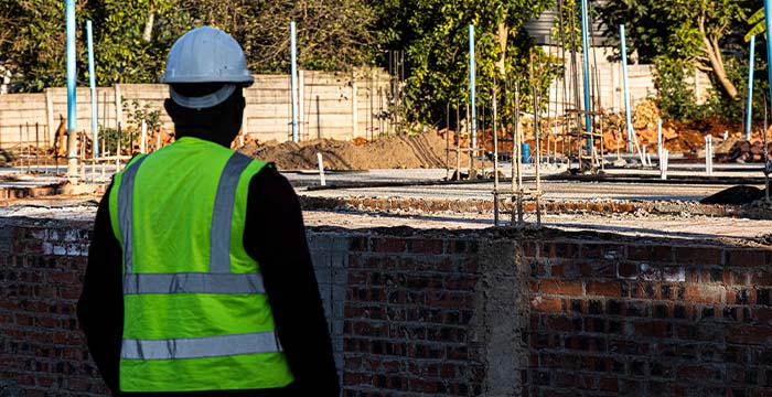 Construction worker in a white helmet and bright green safety vest standing at a worksite with exposed brickwork and foundation structures during daylight.