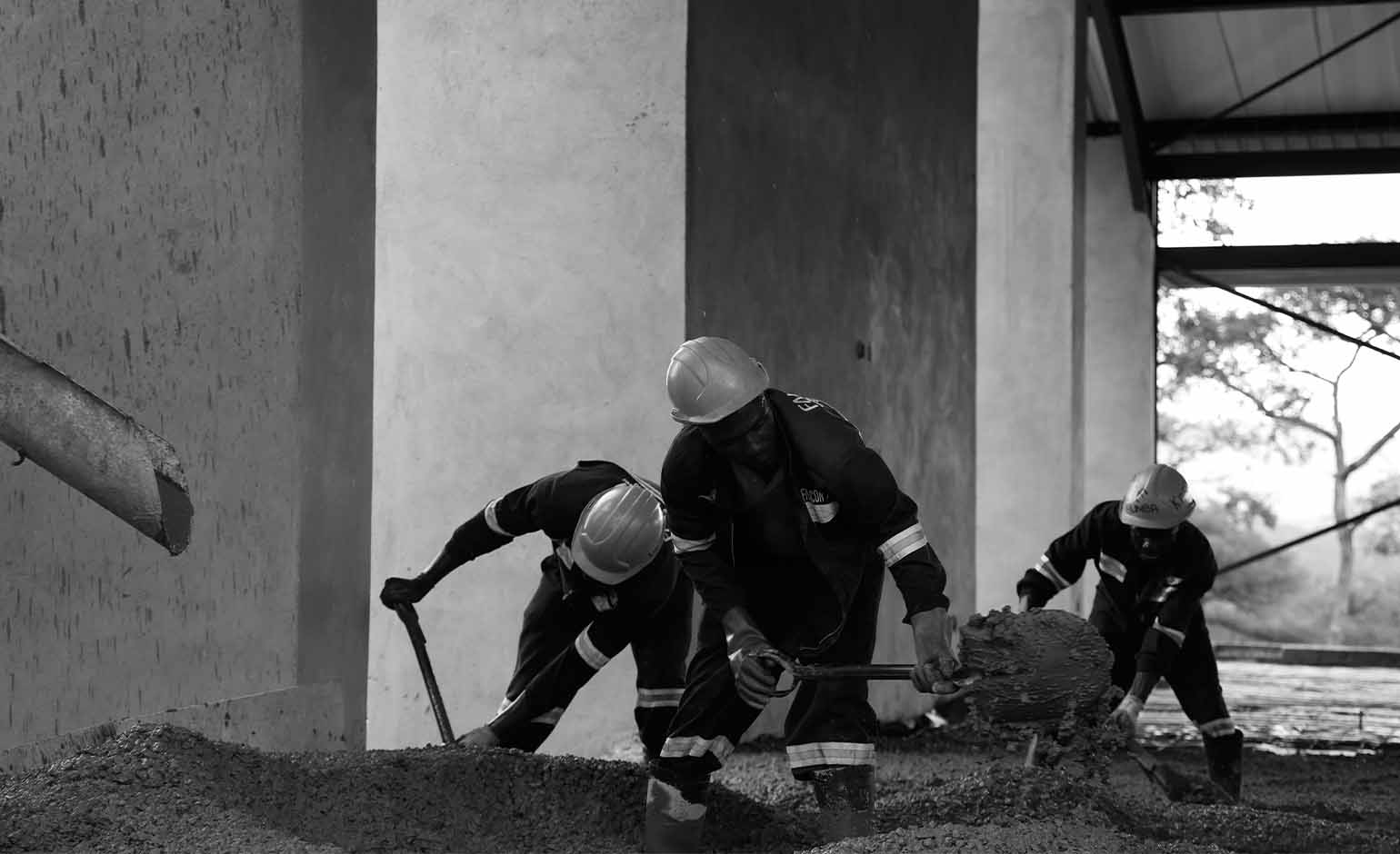 Three construction workers wearing helmets and reflective gear using shovels to smooth and spread concrete inside a building under construction.