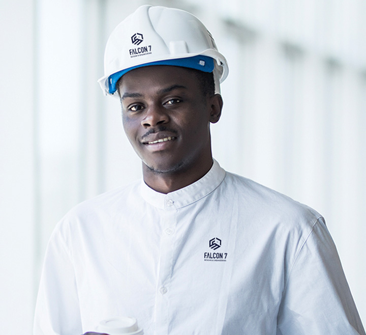 Man wearing a white Falcon 7 branded safety helmet and matching white collared shirt, smiling and holding a cup, with a softly blurred background.