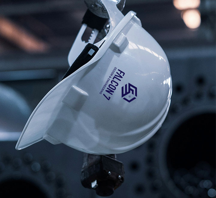 White construction hard hat with the text 'FALCON 7' and a logo, hanging upside down in an industrial setting with blurred machinery in the background.