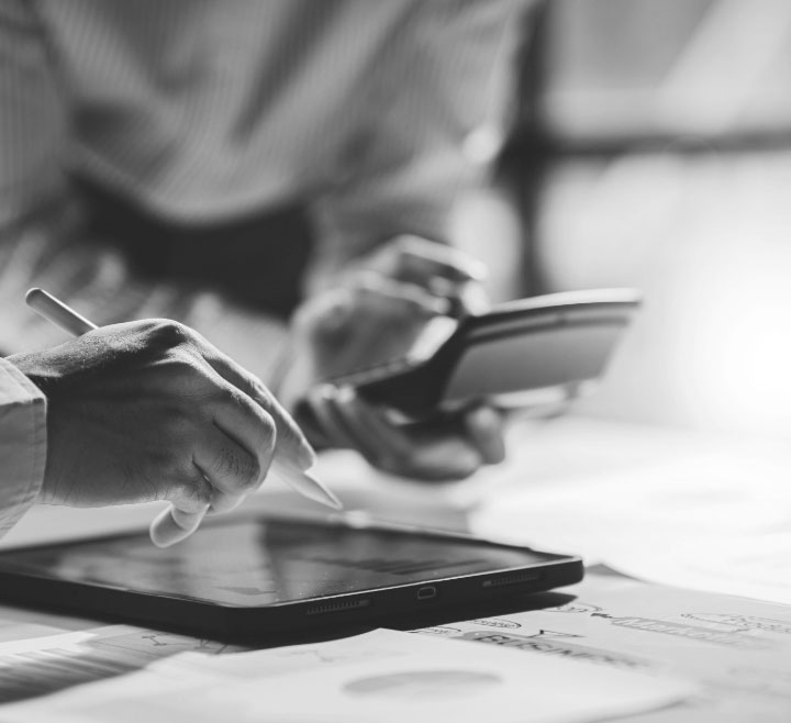 Close-up of hands with a stylus interacting on a tablet while another hand holds a calculator, surrounded by papers with charts and notes.